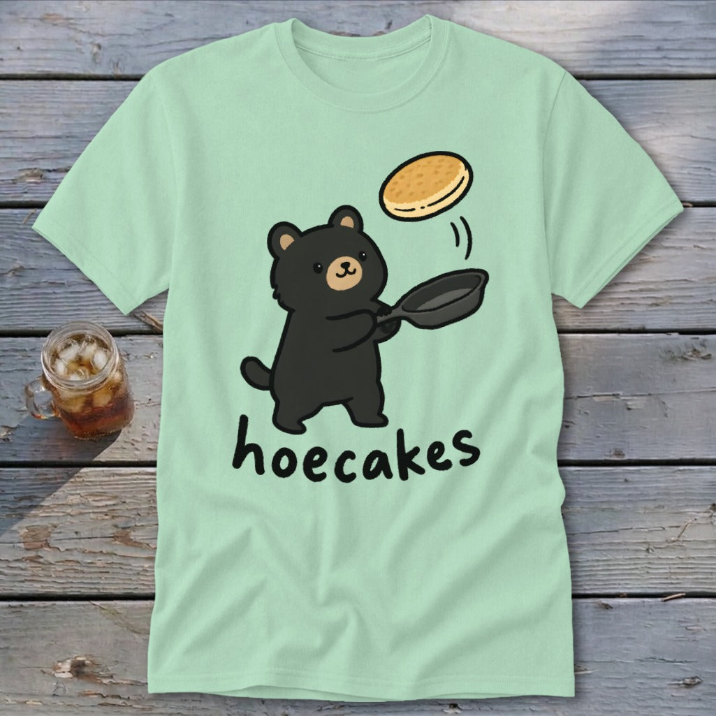 A light green t-shirt featuring a cute black bear flipping a pancake in a pan with the text hoecakes underneath.