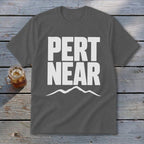 A gray t-shirt with white block text reading PERT NEAR above a mountain range graphic, laid on a wooden surface.