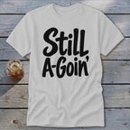 A light grey t-shirt with black text reading "Still A-Goin'" lying flat on a wooden surface.