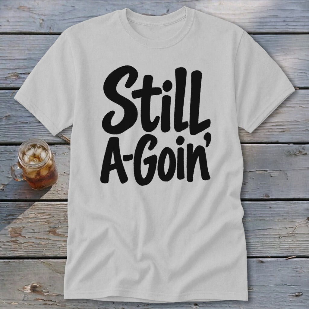 A light grey t-shirt with black text reading "Still A-Goin'" lying flat on a wooden surface.