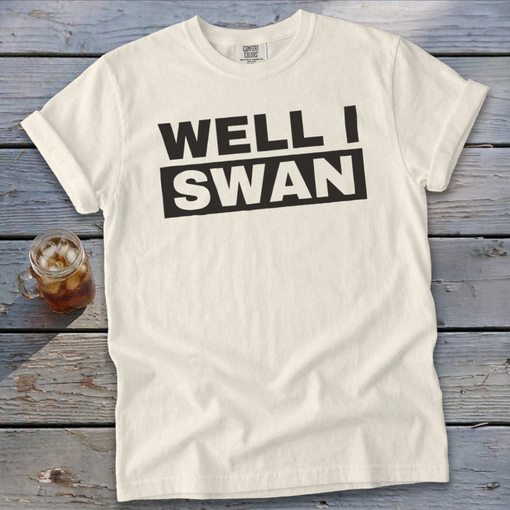 Cream-colored t-shirt with rolled sleeves laid on a wooden surface featuring black text reading WELL I SWAN.