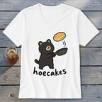 A white V-neck t-shirt featuring a black bear flipping a pancake in a frying pan, with the word hoecakes underneath.