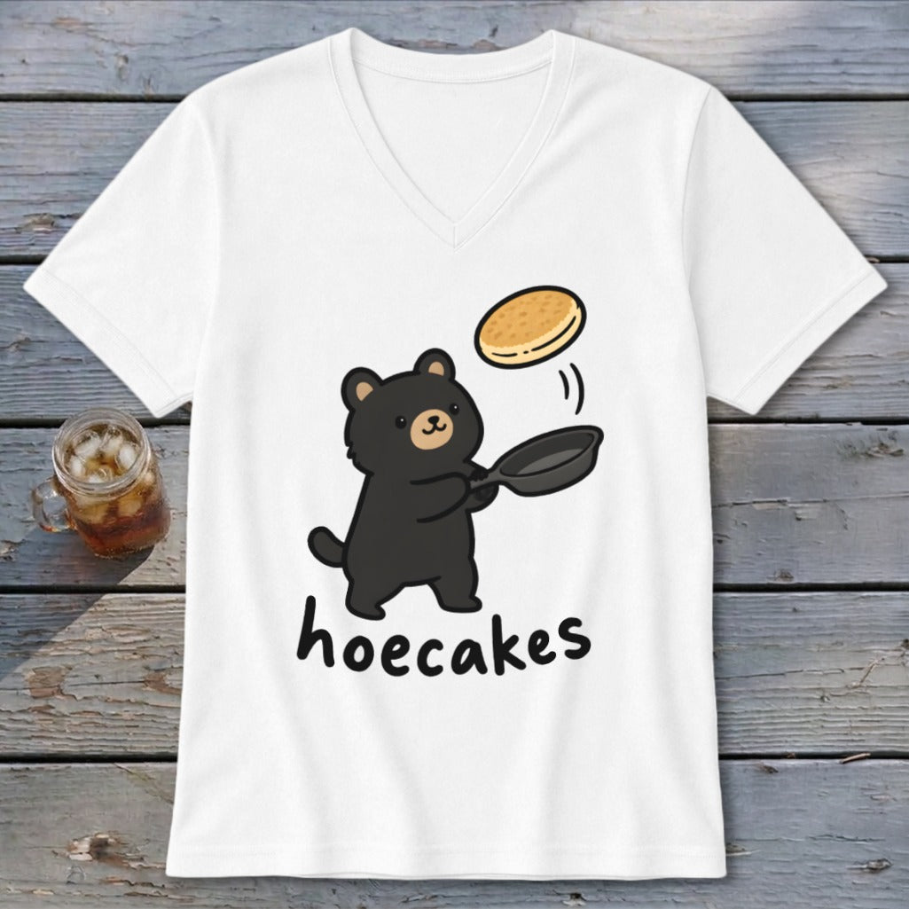 A white V-neck t-shirt featuring a black bear flipping a pancake in a frying pan, with the word hoecakes underneath.
