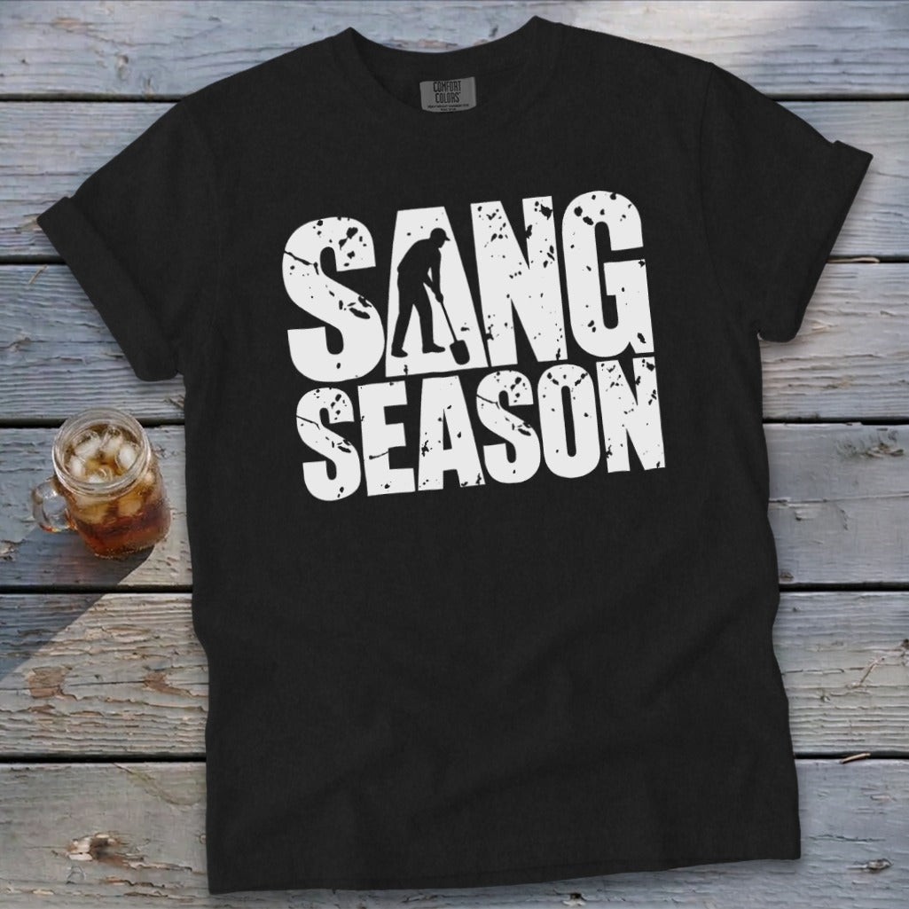 A black short-sleeved t-shirt with distressed white text reading SANG SEASON and a silhouette of a person digging with a shovel inside the letter N, lying on a wooden surface with a glass of iced tea.