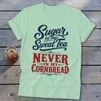 A light green t-shirt with rolled sleeves features blue and red text reading Sugar in my Sweat Tea Never in my Cornbread, laid flat on a rustic wooden background next to a glass of iced tea.