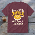 Maroon t-shirt featuring the text Some of Y’all’s Cornbread Ain’t Done in the Middle surrounding a skillet of cornbread.