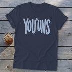 Navy t-shirt with light blue "YOU'UNS" text on a wooden background.