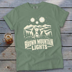 A sage green t-shirt displays white graphics of three people gazing at mountains and glowing lights above.