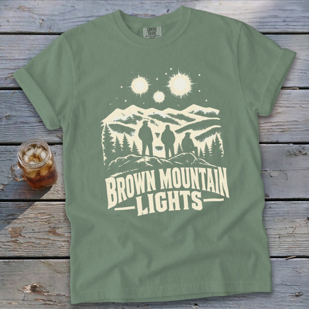 A sage green t-shirt displays white graphics of three people gazing at mountains and glowing lights above.