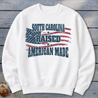 White sweatshirt with distressed American flag graphic and text "SOUTH CAROLINA RAISED AMERICAN MADE".