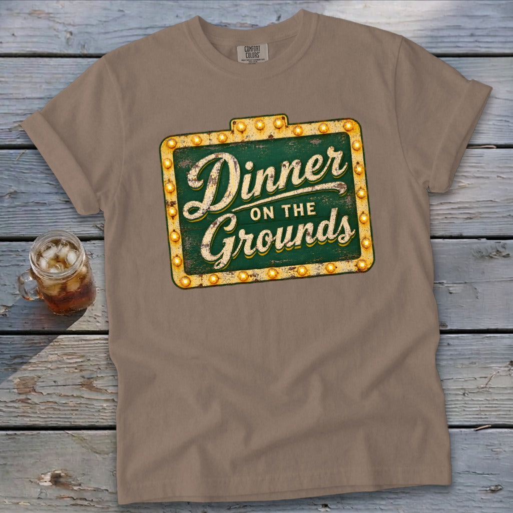 A brown t-shirt featuring a green sign graphic with the text Dinner on the Grounds.
