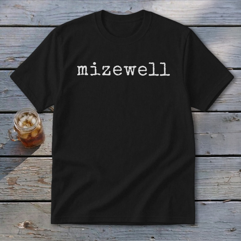Black t-shirt with white text "mizewell" on a wooden surface next to a drink.