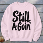 Light pink crewneck sweatshirt featuring the black text "Still A-Goin'" laid flat on a wooden surface.