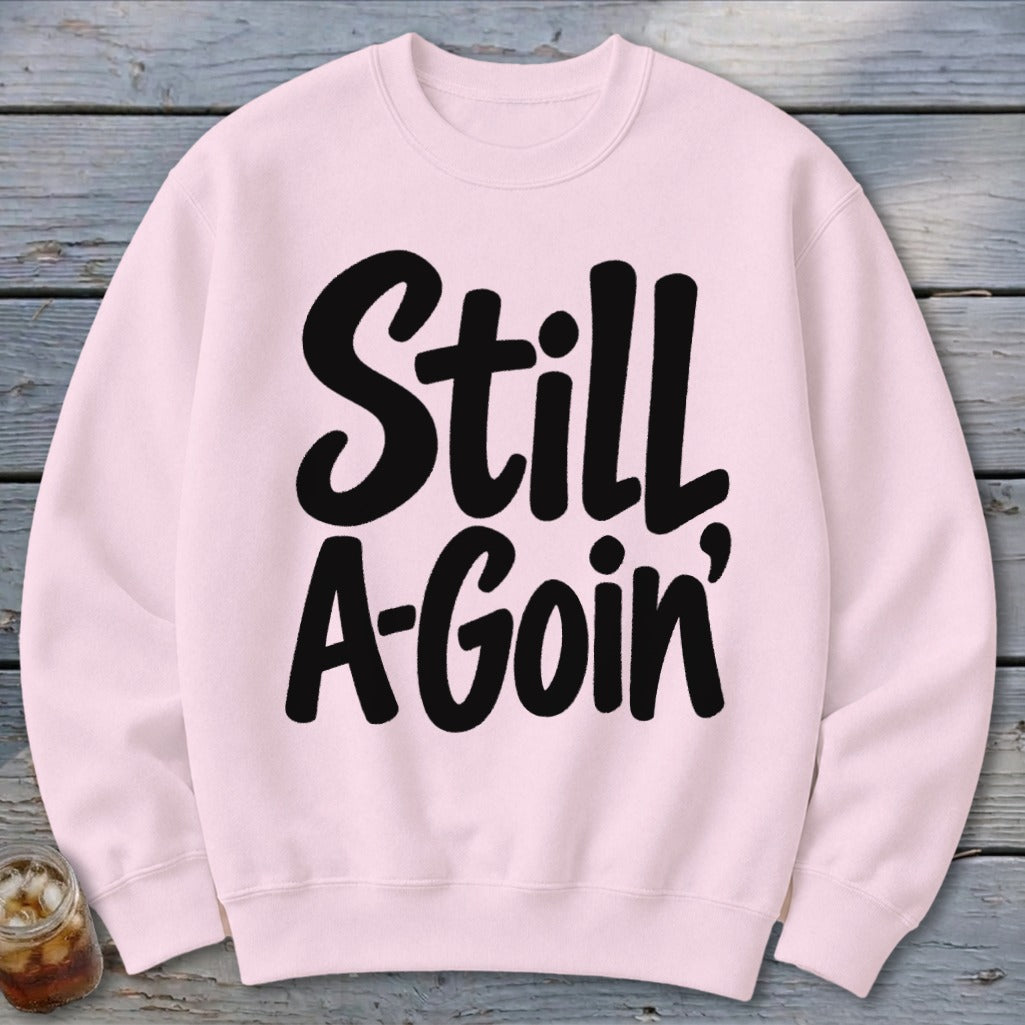 Light pink crewneck sweatshirt featuring the black text "Still A-Goin'" laid flat on a wooden surface.