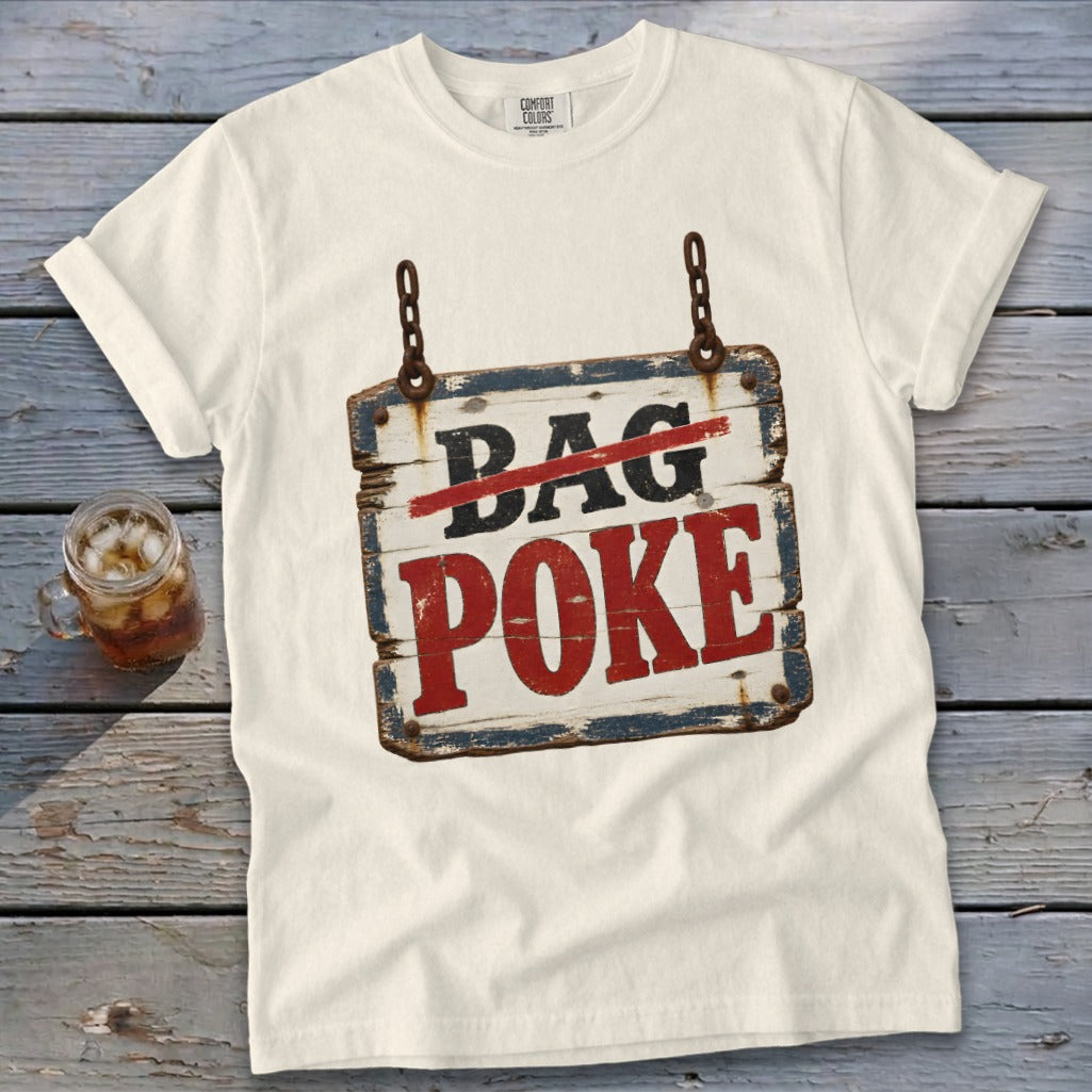 A light cream t-shirt rests on weathered wood, featuring a distressed wooden sign graphic with crossed-out text and red lettering.
