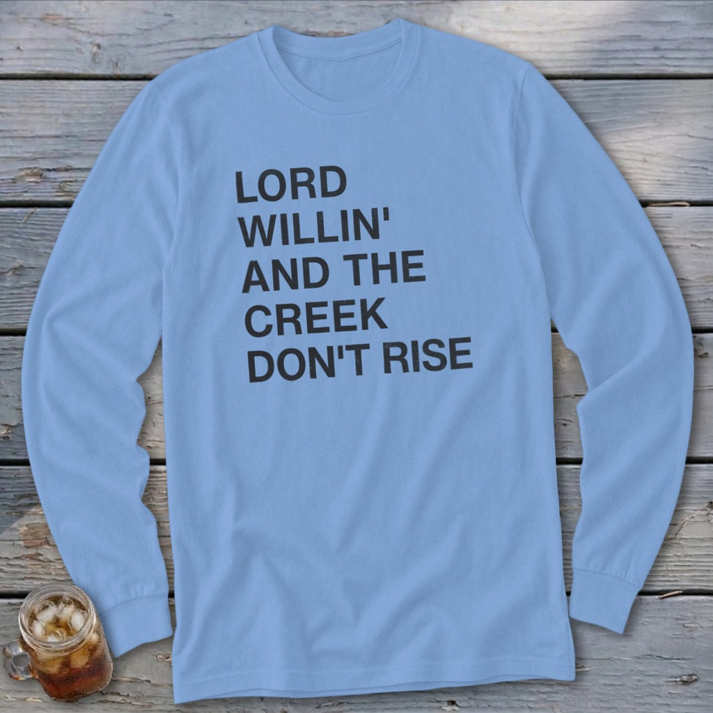 Light blue long-sleeved t-shirt with black text that reads LORD WILLIN' AND THE CREEK DON'T RISE, laid flat on a wooden surface.