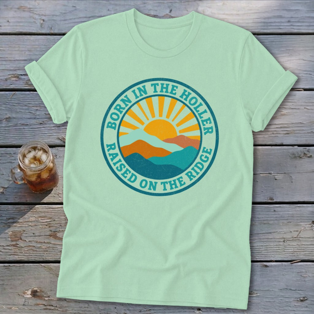 Light green t-shirt featuring a retro graphic of mountains and a sun with the text Born in the Holler Raised on the Ridge.