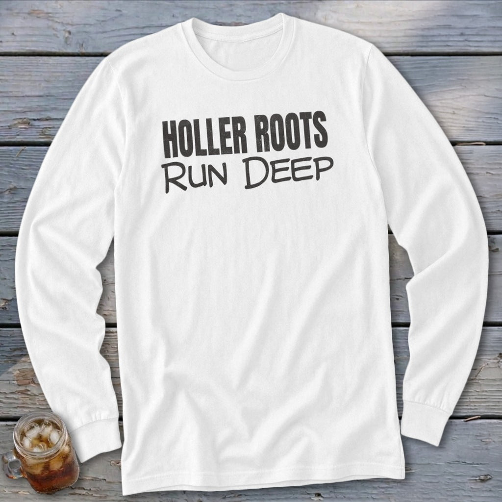 A white long-sleeved t-shirt with black text that reads HOLLER ROOTS RUN DEEP, resting on a rustic wooden background.