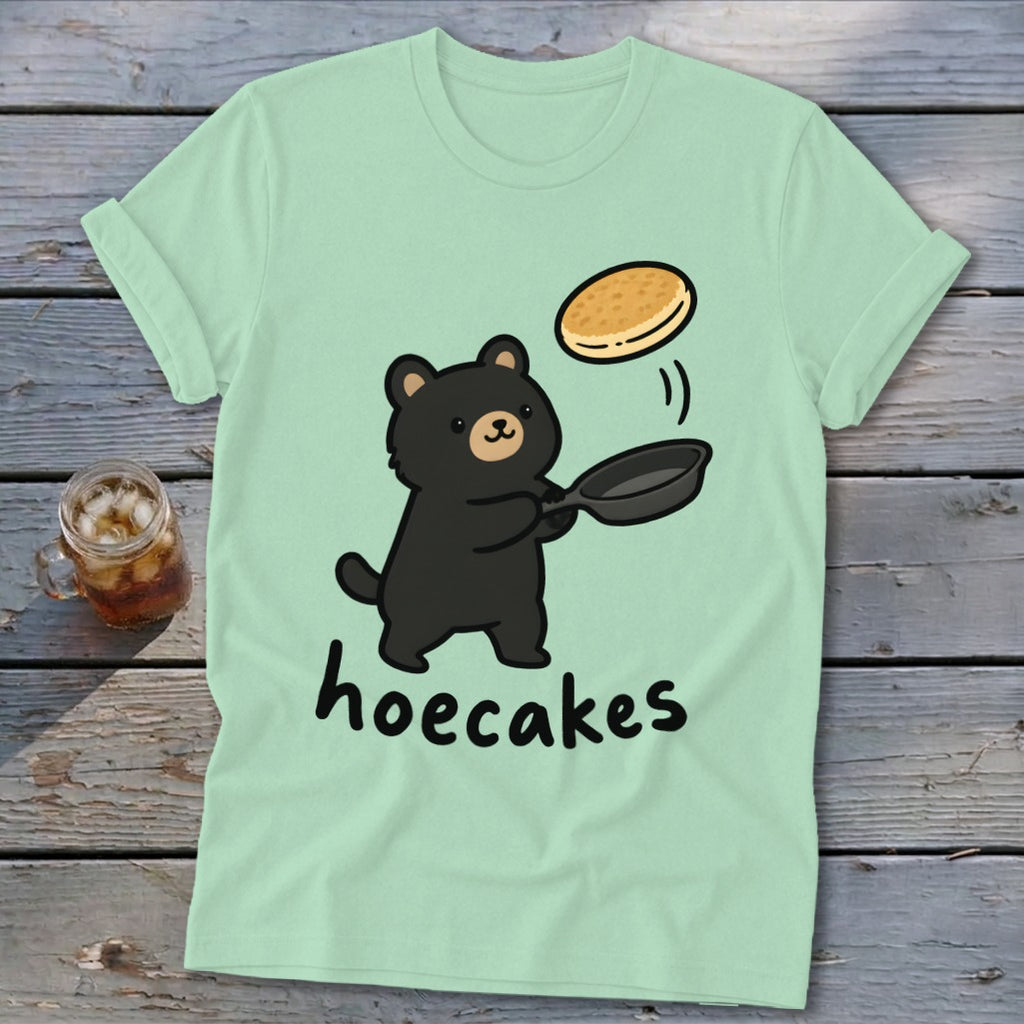 A light green t-shirt featuring a cute black bear flipping a pancake with the text hoecakes.