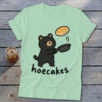 A light green t-shirt featuring a cute black bear flipping a pancake with the text hoecakes.