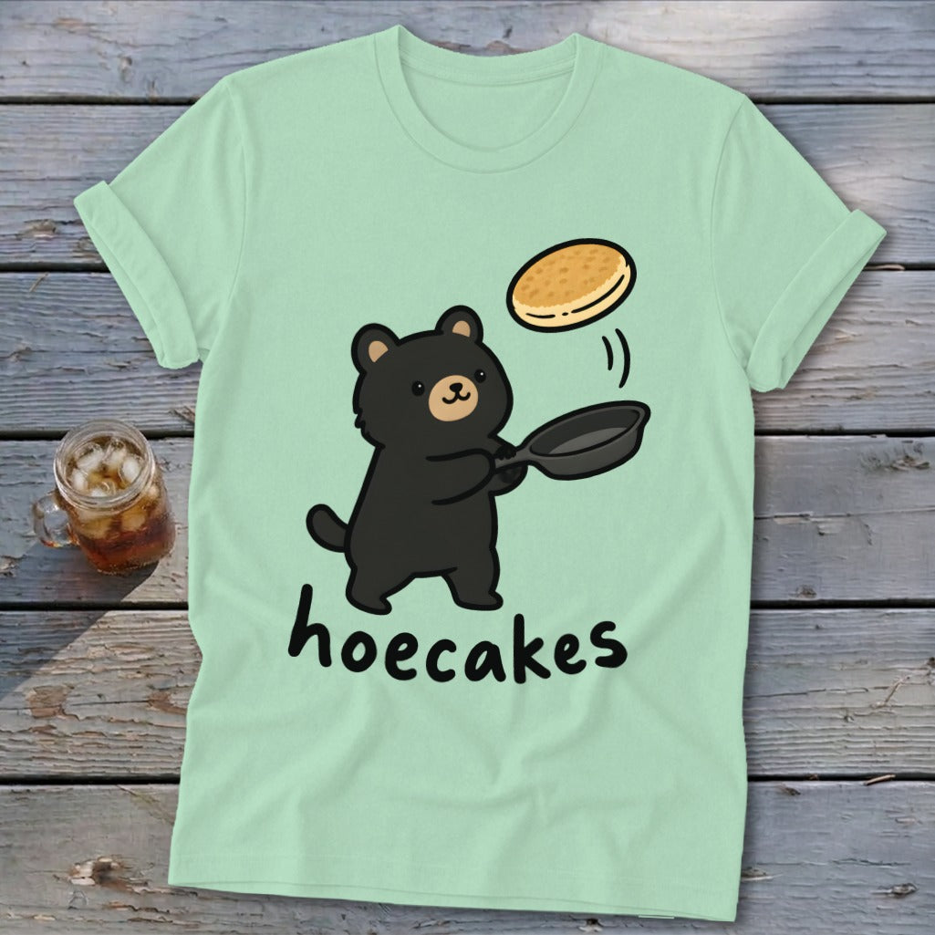 A light green t-shirt featuring a cute black bear flipping a pancake with the text hoecakes.