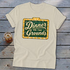 A cream crewneck t-shirt with a vintage green sign graphic reading Dinner on the Grounds.