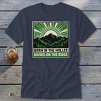 A dark blue t-shirt with a square graphic showing green mountains, a sunburst, and the text BORN IN THE HOLLER RAISED ON THE RIDGE.