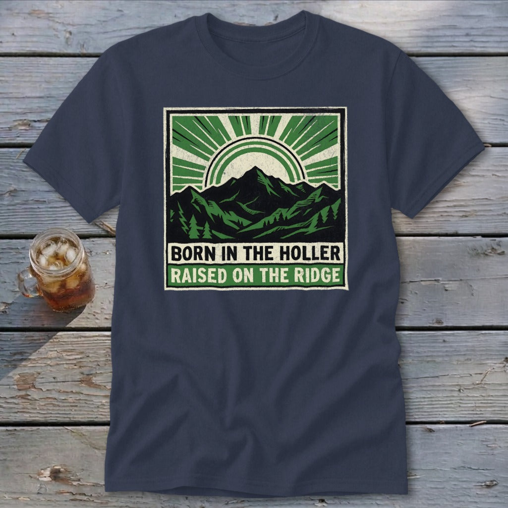 A dark blue t-shirt with a square graphic showing green mountains, a sunburst, and the text BORN IN THE HOLLER RAISED ON THE RIDGE.