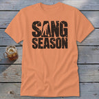 An orange t-shirt featuring distressed black text that reads SANG SEASON with a silhouette of a person digging.