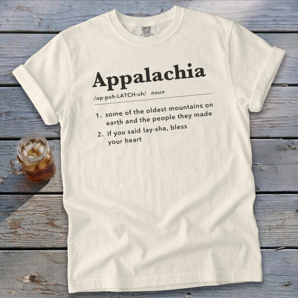 A cream-colored t-shirt with black text defining the word Appalachia rests on a wooden surface beside a glass of iced tea.