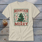 A cream-colored short-sleeve t-shirt lies on a wooden surface, featuring a retro Christmas tree graphic with the text MOUNTAIN MERRY.