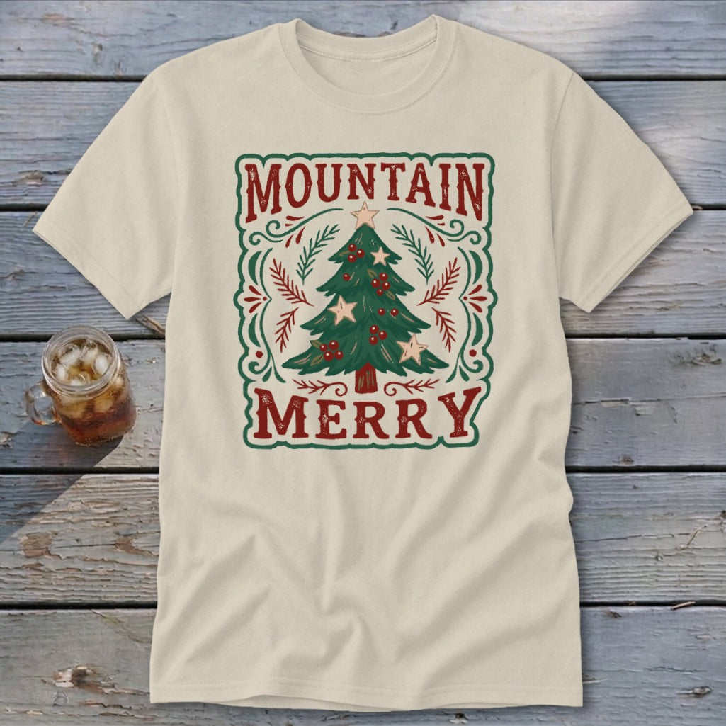 A cream-colored short-sleeve t-shirt lies on a wooden surface, featuring a retro Christmas tree graphic with the text MOUNTAIN MERRY.