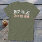 Olive green t-shirt with white and pink text reading "THESE HOLLERS KNOW MY NAME".