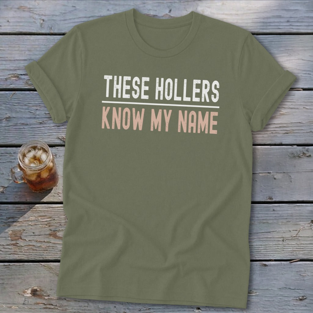 Olive green t-shirt with white and pink text reading "THESE HOLLERS KNOW MY NAME".