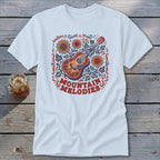 A light blue t-shirt featuring a folk art design of a guitar, sunflowers, leaves, and the text "MOUNTAIN MELODIES" on a wooden surface.