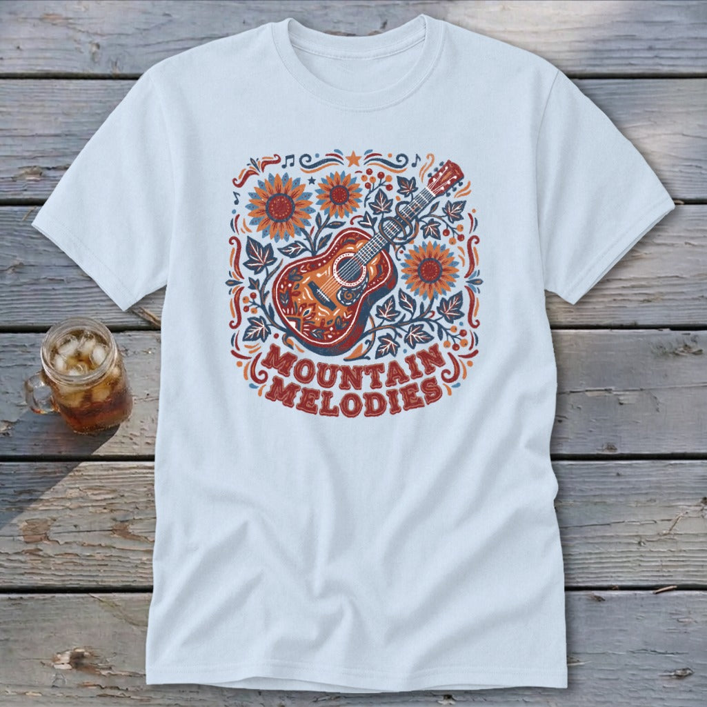 A light blue t-shirt featuring a folk art design of a guitar, sunflowers, leaves, and the text "MOUNTAIN MELODIES" on a wooden surface.
