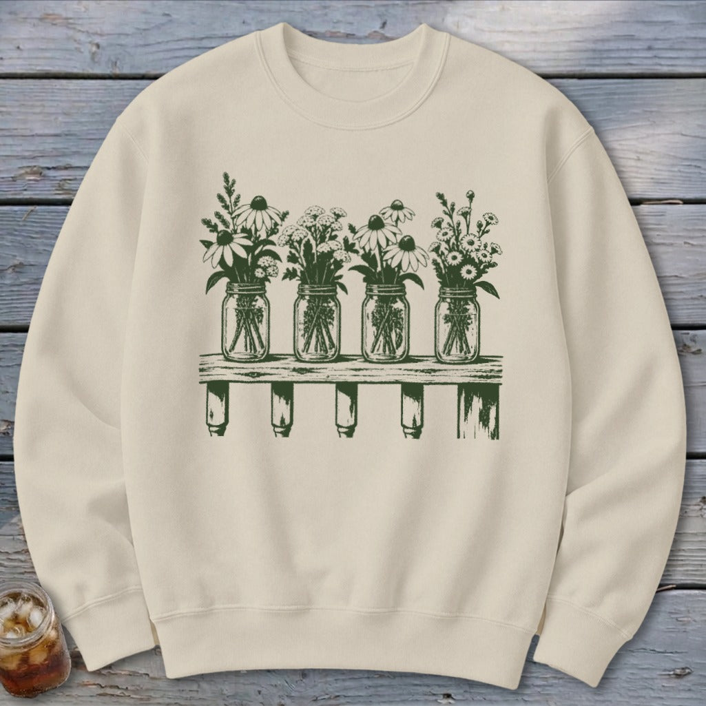 Beige crewneck sweatshirt with a green graphic of four mason jars filled with wildflowers on a wooden shelf.