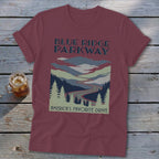 Maroon t-shirt with a vintage Blue Ridge Parkway landscape graphic and text on a wooden surface.