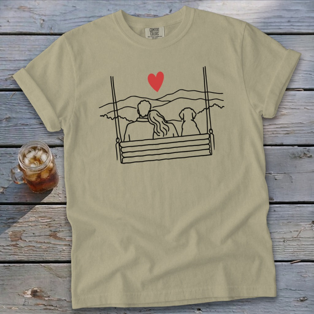 Khaki t-shirt with a line drawing of a couple and dog on a swing above mountains.