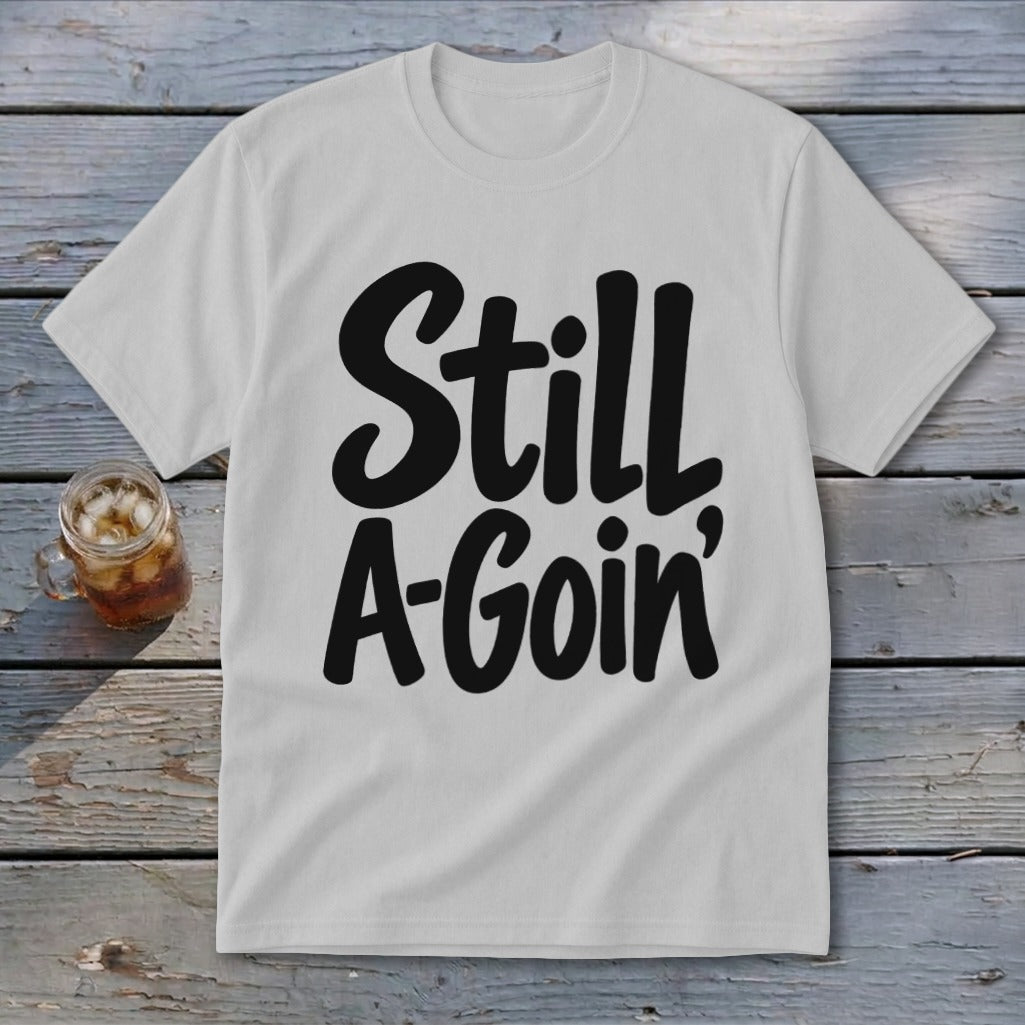 Light gray crewneck t-shirt featuring the black text Still A-Goin centered on the chest, laid flat on a rustic wooden surface next to a small glass jar filled with iced tea.