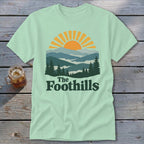 Light green t-shirt with a mountain sunset graphic and "The Foothills" text.