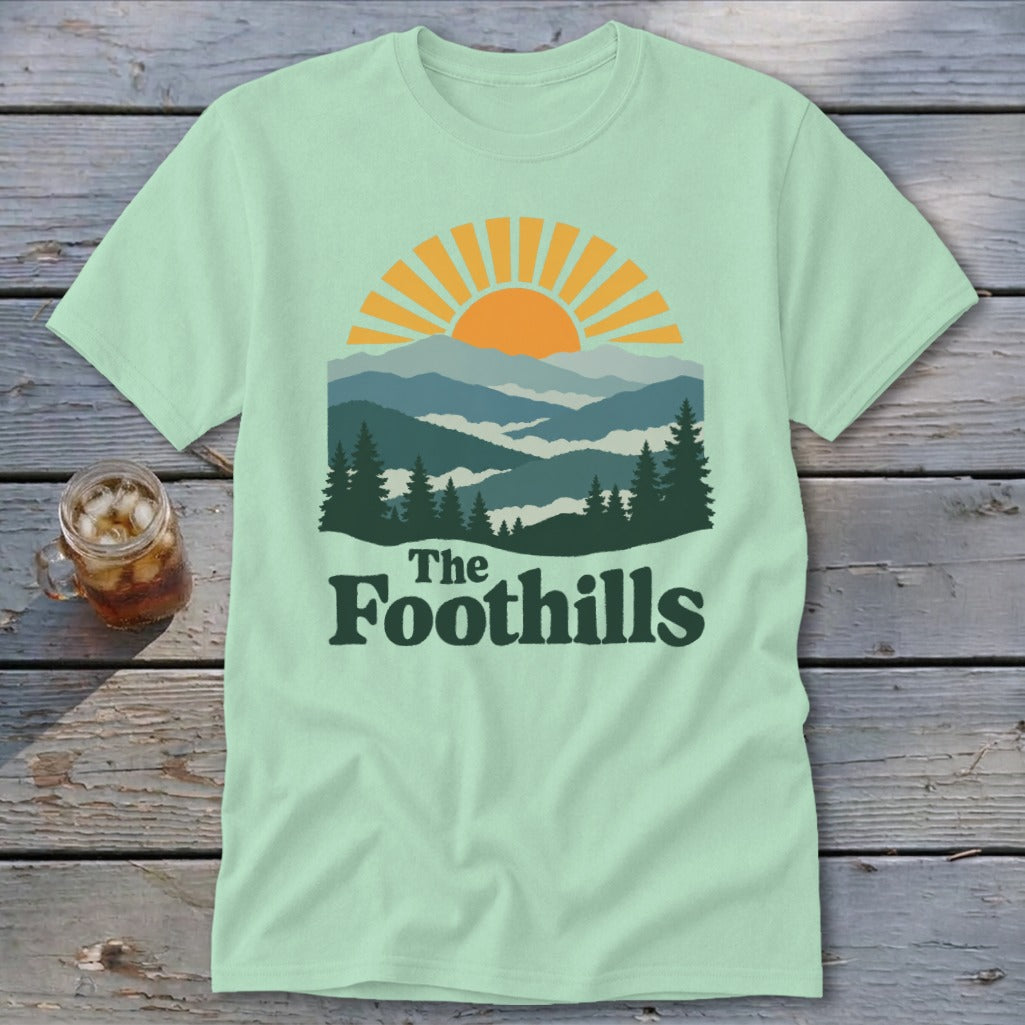 Light green t-shirt with a mountain sunset graphic and "The Foothills" text.