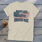 Cream-colored crewneck t-shirt with rolled sleeves laid flat on a rustic wooden surface, featuring distressed text and a US flag graphic that reads "Maryland Raised American Made."