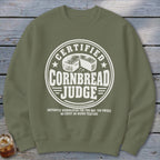 An olive green crewneck sweatshirt featuring a circular white graphic with the text Certified Cornbread Judge and a cornbread illustration.