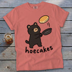 A salmon pink t-shirt features a cartoon black bear flipping a pancake from a frying pan above the word hoecakes.