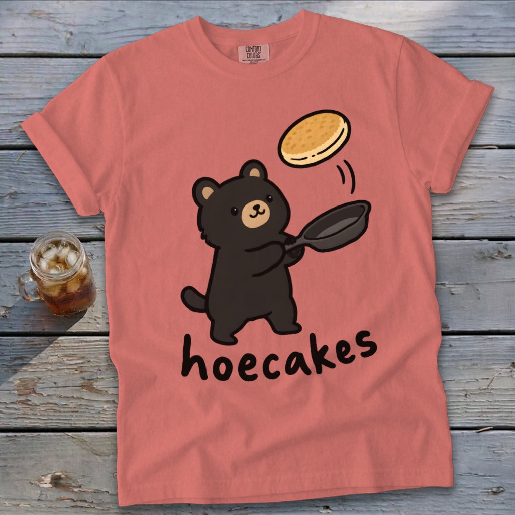A salmon pink t-shirt features a cartoon black bear flipping a pancake from a frying pan above the word hoecakes.
