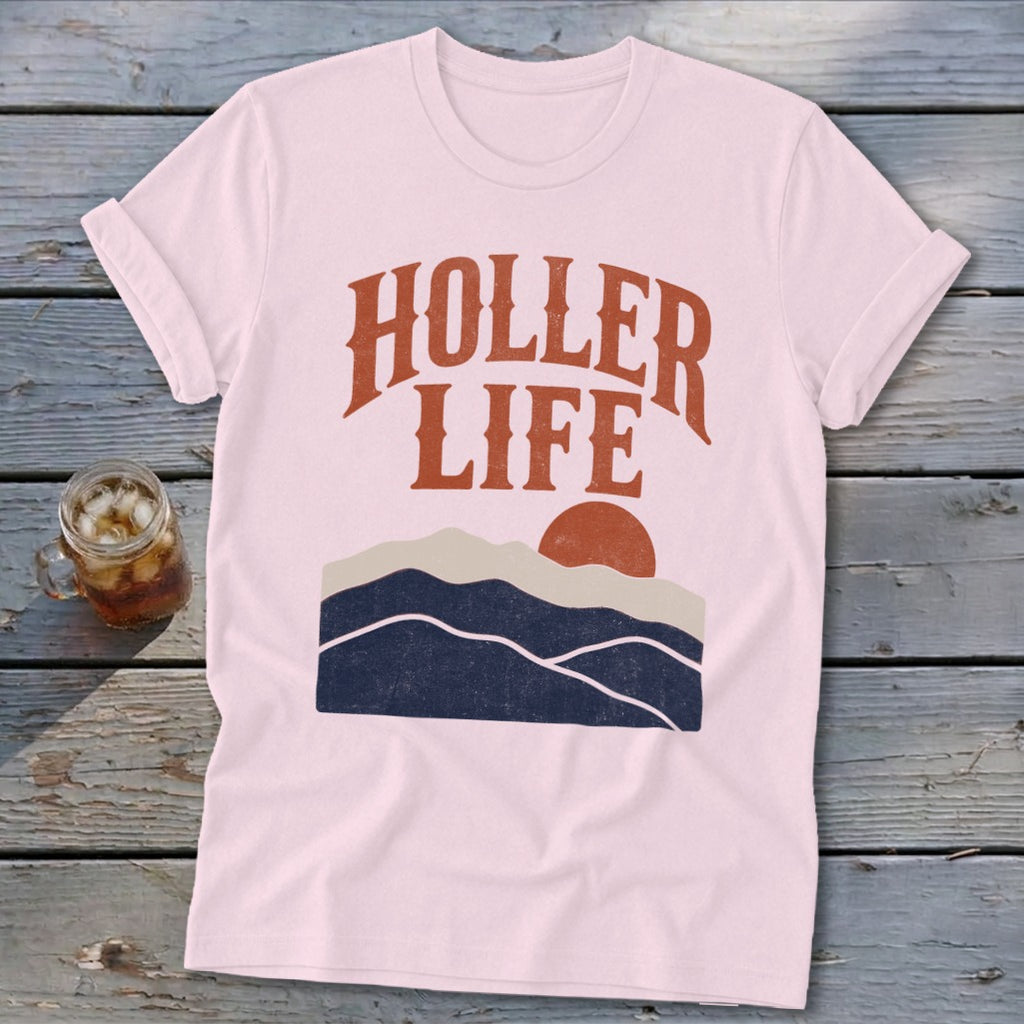 Light pink short-sleeve t-shirt with orange text reading HOLLER LIFE above a sunset mountain graphic, laid on weathered wood.