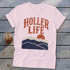 Light pink short-sleeve t-shirt with orange text reading HOLLER LIFE above a sunset mountain graphic, laid on weathered wood.