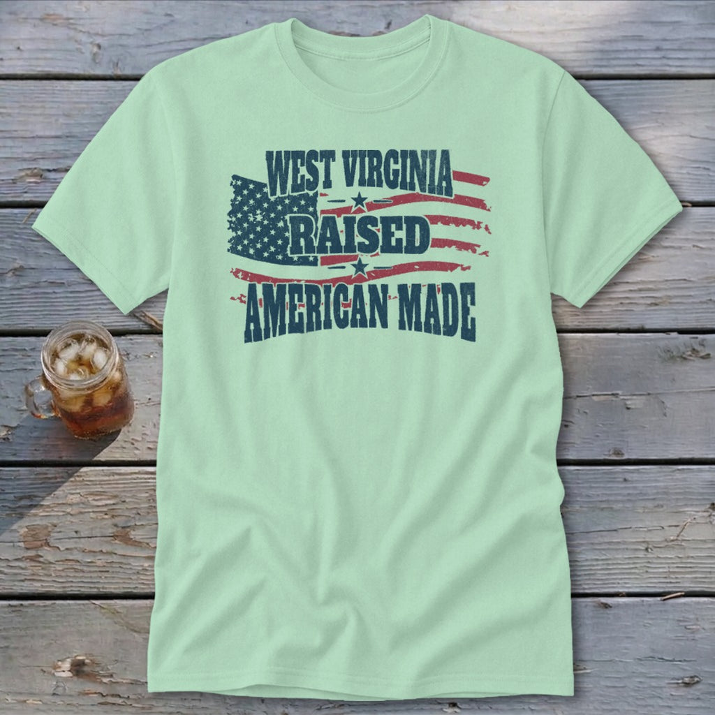 A light green t-shirt featuring a distressed American flag graphic with the text West Virginia Raised American Made.