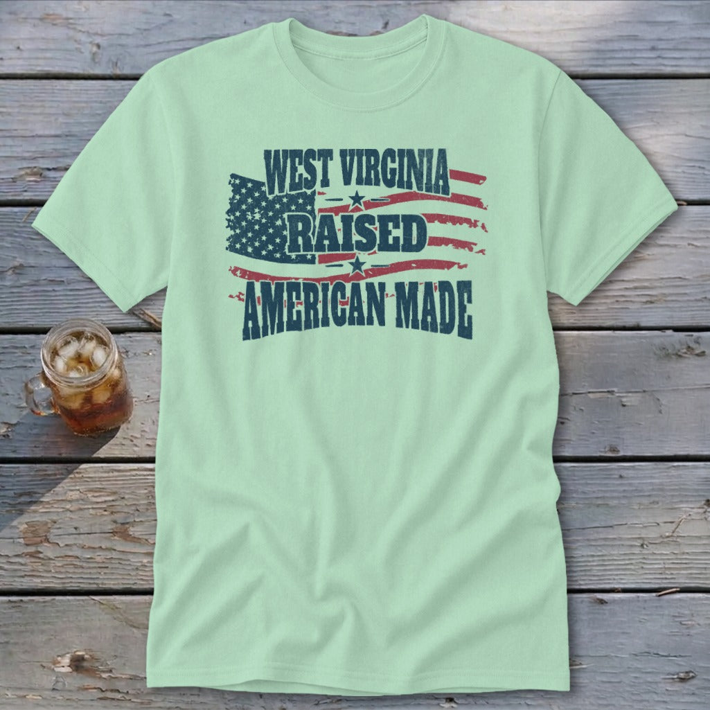 A light green t-shirt featuring a distressed American flag graphic with the text West Virginia Raised American Made.